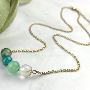 Może przedstawiać: Złoty łańcuszek z rzędem okrągłych zielonych, niebieskich i przezroczystych koralik&oacute;w.