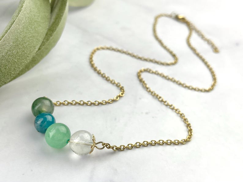 Może przedstawiać: Złoty łańcuszek z rzędem okrągłych zielonych, niebieskich i przezroczystych koralik&oacute;w.