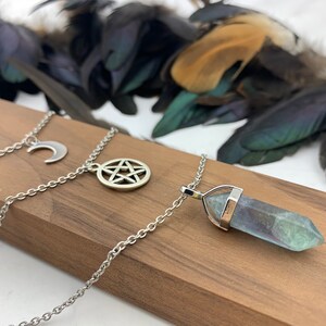 Pu&ograve; includere: Collane argentate con ciondoli celesti e spirituali. Una collana presenta una luna crescente, un'altra un pentacolo e la terza un ciondolo di cristallo blu e verde. Le collane sono appoggiate su una superficie di legno.