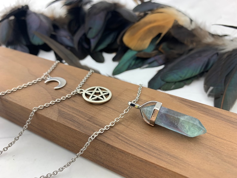 Pu&ograve; includere: Collane argentate con ciondoli celesti e spirituali. Una collana presenta una luna crescente, un'altra un pentacolo e la terza un ciondolo di cristallo blu e verde. Le collane sono appoggiate su una superficie di legno.