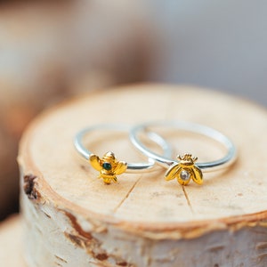 Pode incluir: Dois an&eacute;is de prata com motivos de abelhas em tom dourado. Uma abelha tem uma pequena pedra verde-escura no centro, enquanto a outra apresenta uma pedra transparente e brilhante. Os aros s&atilde;o finos, polidos e os desenhos s&atilde;o detalhados.