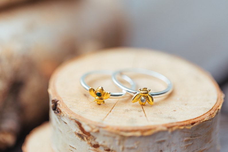 Pode incluir: Dois an&eacute;is de prata com motivos de abelhas em tom dourado. Uma abelha tem uma pequena pedra verde-escura no centro, enquanto a outra apresenta uma pedra transparente e brilhante. Os aros s&atilde;o finos, polidos e os desenhos s&atilde;o detalhados.