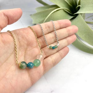 Może przedstawiać: Dwa naszyjniki z zielonymi i niebieskimi koralikami z kamieni szlachetnych na złotym i srebrnym łańcuszku. Naszyjniki są prezentowane na dłoni.