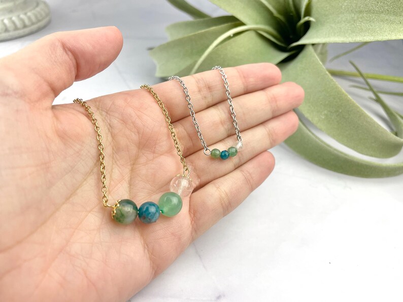 Może przedstawiać: Dwa naszyjniki z zielonymi i niebieskimi koralikami z kamieni szlachetnych na złotym i srebrnym łańcuszku. Naszyjniki są prezentowane na dłoni.