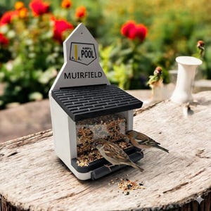 May include: A white and black bird feeder with the text "ALBERTA POOL MUIRFIELD" is filled with birdseed. Two small birds are perched on the feeder, with scattered seeds on the wooden surface. The feeder has a black roof.