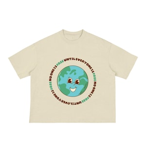 May include: A cream-coloured t-shirt with a smiling Earth graphic. The design includes the text "EVERYONE IS FREE UNTIL NO ONE IS FREE" in a circular pattern. The Earth graphic is in shades of blue and green, with a friendly face.