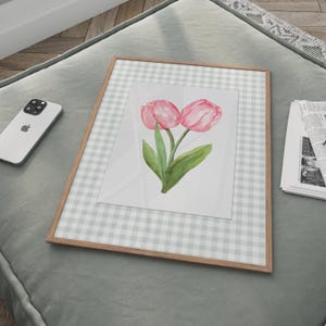 May include: A framed watercolor print featuring two pink tulips with green leaves. The artwork is set against a light gray and white gingham background and framed in a light brown wood. The print is displayed on a gray surface.