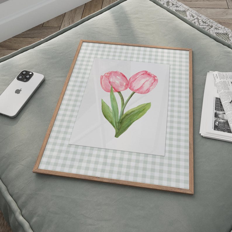 May include: A framed watercolor print featuring two pink tulips with green leaves. The artwork is set against a light gray and white gingham background and framed in a light brown wood. The print is displayed on a gray surface.