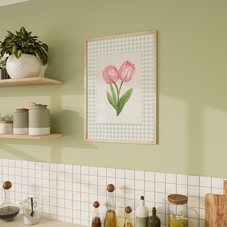May include: A framed watercolor print of two pink tulips with green leaves. The artwork is set against a light green and white gingham pattern and framed in light wood. The print hangs on a light green wall.