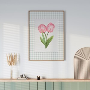May include: A framed watercolor painting of two pink tulips with green leaves. The artwork is set against a light green and white gingham background and framed in a light brown wooden frame. The painting is displayed on a shelf with other decorative items.