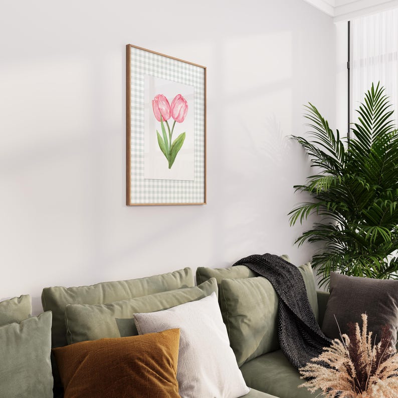 May include: A framed watercolor print featuring two pink tulips with green leaves. The artwork is set against a white and light gray gingham pattern, encased in a thin, gold-colored frame. The print is displayed on a white wall above a green sofa with various pillows.