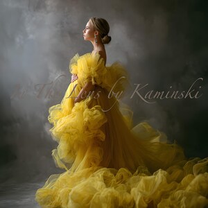 May include: A woman in a vibrant yellow gown with a ruffled design and a long train. The dress is strapless and set against a smoky grey backdrop. The text "In The Lens by Kaminski" is visible on the image.