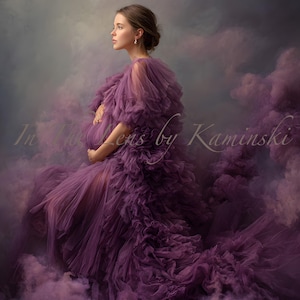 May include: A woman in a long, ruffled purple dress, set against a cloudy background. The dress features a layered design and a flowing train. The image has soft lighting and a dreamy feel. The text "In the lens by Kaminski" is visible.