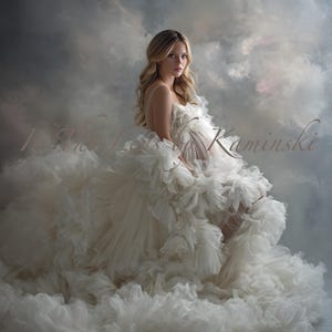 Puede incluir: Una mujer con un vestido de maternidad blanco roto con volantes en capas y una larga cola. El vestido tiene tirantes finos. El fondo es un cielo nublado. La imagen incluye el texto "In Anna Lisa Kaminski" en cursiva.