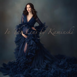 Puede incluir: Un vestido de maternidad azul marino con escote pronunciado y mangas largas. El vestido presenta capas de tela con volantes y una abertura en el muslo. El texto "In The Lens by Kaminski" es visible.