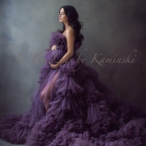 Puede incluir: Un vestido de maternidad morado, sin tirantes, largo hasta el suelo, con un diseño de volantes en capas. El vestido presenta una abertura alta y una larga cola. La imagen incluye el texto "In The Lens by Kaminski".