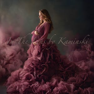 Puede incluir: Una mujer con un vestido de maternidad fluido de color ciruela. El vestido presenta un corpiño plisado y una falda voluminosa con volantes. La imagen tiene una calidad suave y etérea con un fondo ahumado y rosado. El texto en la imagen dice "In The Lens by Kaminski".