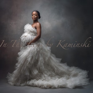 May include: A woman in a white, strapless, layered gown with a long train. The gown has a ruffled bodice and skirt. The background is a muted grey with the text "In The Lens by Kaminski" in a soft, cursive font.