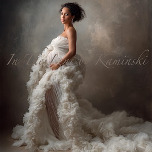 May include: A woman in a white, strapless maternity gown with a layered, ruffled skirt and a long train. The gown has a flowing, ethereal quality. The image includes the text "In The Lens by Kaminski".