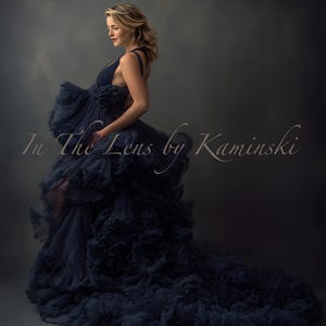 Puede incluir: Una mujer con un vestido azul marino fluido con una falda con volantes en capas y una larga cola. El vestido tiene un cuello halter y se usa en un estudio. El texto "In The Lens by Kaminski" es visible.