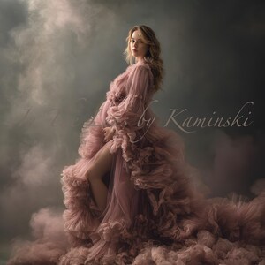 Puede incluir: Una mujer con un vestido de maternidad rosa polvoriento y fluido. El vestido presenta capas de tela con volantes y mangas largas. La imagen tiene una calidad suave y etérea con un fondo ahumado. El texto "by Kaminski" es visible.
