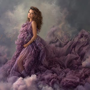 Puede incluir: Una mujer con un vestido de maternidad lavanda fluido. El vestido presenta capas de tela con volantes y una larga cola, sobre un fondo de texturas suaves y nubosas. La imagen incluye el texto "In The Lens Kaminski".