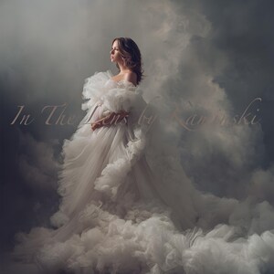 May include: A woman in a white, layered, ruffled gown with a long train. The off-the-shoulder dress is set against a backdrop of soft, cloudy textures. The text "In The Lens by Kaminski" is visible, adding a delicate touch to the image.
