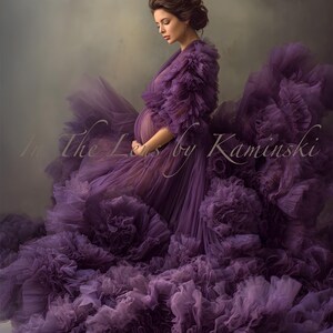 Puede incluir: Una mujer con un vestido morado fluido y con volantes. El vestido presenta capas de tela y un escote con volantes. La imagen tiene el texto "In The Lens by Kaminski" en la esquina inferior izquierda. El fondo es gris suave y apagado.