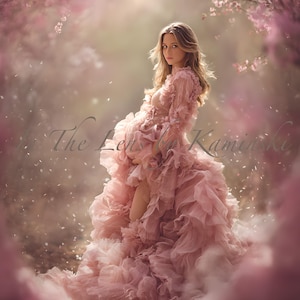 May include: A woman in a flowing, pale pink gown with ruffled layers stands in a soft, dreamy outdoor setting. The dress has long sleeves and a high neckline. The background features soft pink blossoms and the text "The Lens by Kaminski".
