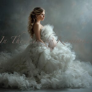 May include: A woman wearing a white, ruffled maternity gown. The dress features a strapless design and a full skirt. The woman has fair skin and blonde hair, looking to the side. The image includes the text "In The Lens by Kaminski".
