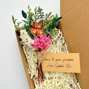 Op de afbeelding: Een kleine boeket gedroogde bloemen in een kartonnen doos. Het boeket bevat roze, oranje en witte bloemen, met groen blad. Een klein kaartje leest "Cheers to your freedom Love, Sophie Xx". De doos is gevuld met papier.