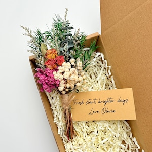 May include: A small bouquet of dried flowers in a cardboard box. The bouquet includes orange roses, pink and white flowers, and green foliage, tied with twine. A small card reads "Fresh start, brighter days, Love, Olivia."