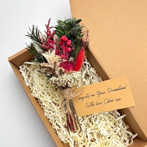 May include: A small bouquet of dried flowers in a cardboard box with shredded paper. The bouquet features red, white, and green elements, tied with twine. A card reads "Congrats on Your Promotion! With love, Chloe xxx."