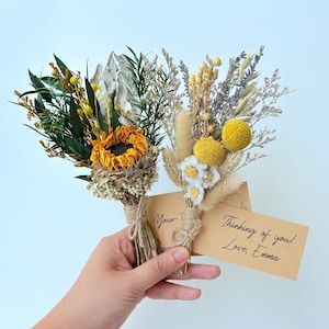 Op de afbeelding: Twee kleine boeketten van gedroogde bloemen met zonnebloemen, gele, witte en lavendelkleurige bloemen, vastgebonden met touw. Elk boeket heeft een klein kraftpapier kaartje met "Thinking of you! Love, Emma."