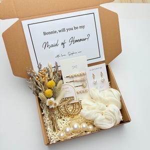 May include: A brown gift box containing a card with the text "Bonnie, will you be my Maid of Honour?" Gold hair clips, pearl earrings, a dried flower bouquet, and a cream scrunchie are also inside. The box is filled with paper.