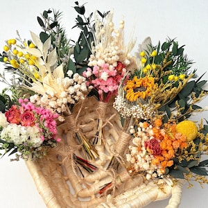 May include: A collection of dried flower bouquets in a woven basket. The bouquets feature a variety of colors, including yellow, pink, orange, and white. Each bouquet is tied with twine. The basket is beige.