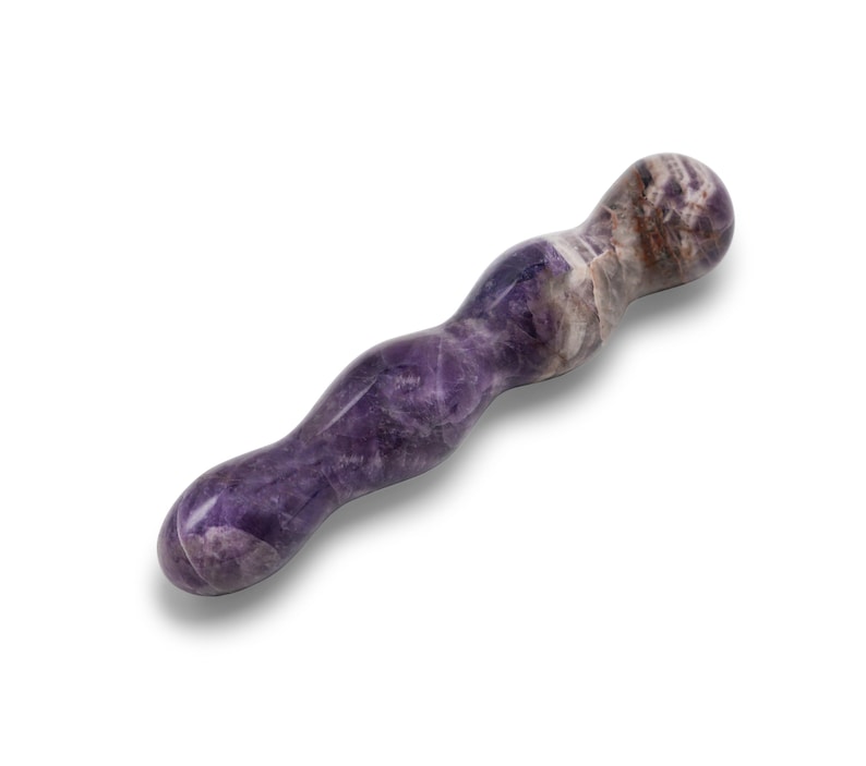 May include: A purple and white amethyst wand with a curved, ergonomic design. The polished stone features natural colour variations and a smooth, rounded tip. The wand is designed for massage or therapeutic use.