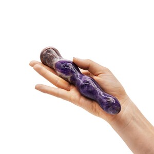 May include: A purple and white amethyst crystal wand. The wand is held in a hand against a white background. The crystal has a smooth, polished surface and a rounded tip.