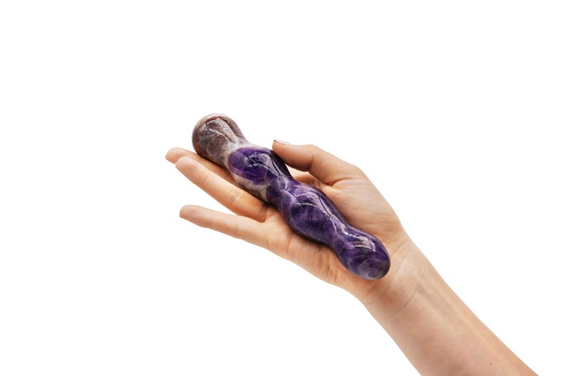 May include: A purple and white amethyst crystal wand. The wand is held in a hand against a white background. The crystal has a smooth, polished surface and a rounded tip.