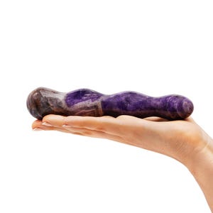 May include: A purple and brown stone sex toy with a wavy design. The product is held in a hand against a white background. The stone has a polished finish and a natural, organic appearance.