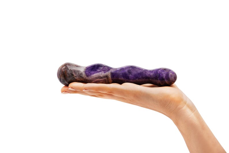 May include: A purple and brown stone sex toy with a wavy design. The product is held in a hand against a white background. The stone has a polished finish and a natural, organic appearance.