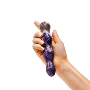 May include: A purple and brown amethyst crystal wand. The wand is held in a hand against a white background. The wand has a smooth, polished surface and a rounded shape.
