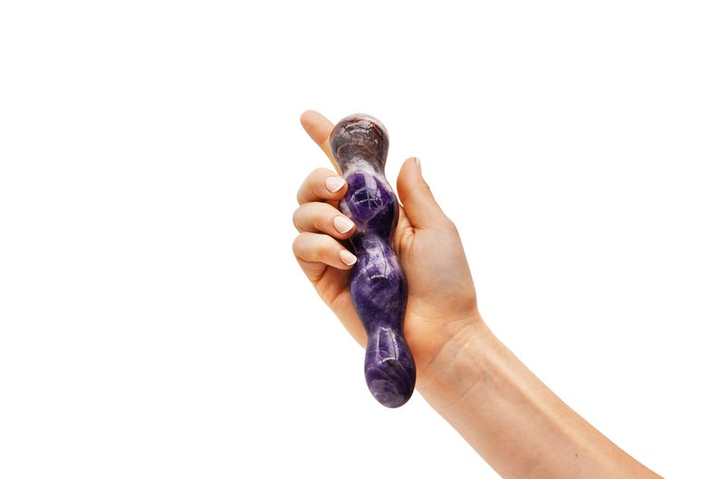 May include: A purple and brown amethyst crystal wand. The wand is held in a hand against a white background. The wand has a smooth, polished surface and a rounded shape.