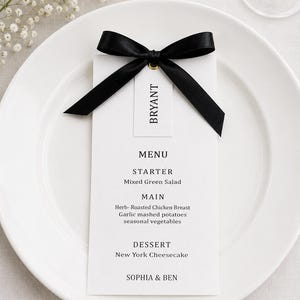 May include: A white rectangular menu card with black text reading MENU, STARTER Mixed Green Salad, MAIN Herb-Roasted Chicken Breast, Garlic mashed potatoes, seasonal vegetables, DESSERT New York Cheesecake, SOPHIA & BEN, topped with a black satin ribbon and a name tag.