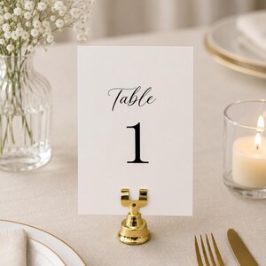 May include: A white rectangular table number card with the black text Table 1 printed in a serif font. The card is held upright by a small, polished gold-colored metal stand with a U-shaped clip at the top and a circular base.