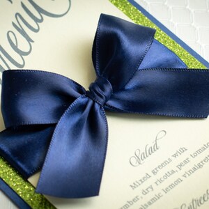 May include: A green and blue wedding menu card with a navy blue satin bow on top. The menu card has the word "Menu" written in cursive at the top and the word "Salad" written in cursive below the bow. The salad description is "Mixed greens with amber, dry ricotta, pear tomato and balsamic lemon vinaigrette".