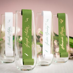 May include: Four champagne flutes with green and white place cards. The place cards are rolled and tied with a ribbon. The names on the place cards are Madeline Allen, Joseph Collier, Justin Young, and Amanda Diaz.