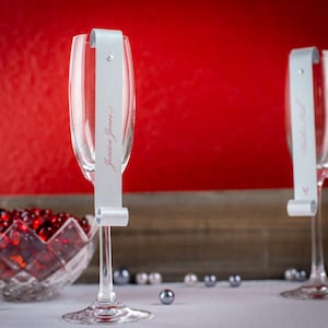 May include: Two champagne flutes with silver place cards. The place cards are attached to the stems of the glasses with a small hole. The place card on the left glass reads "Jessica Jones".