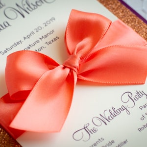 May include: A coral satin ribbon tied into a bow on a white wedding invitation with gold glitter trim. The invitation text reads "The Wedding Party" and lists the parents of the groom, Robert & Susan Wilson.