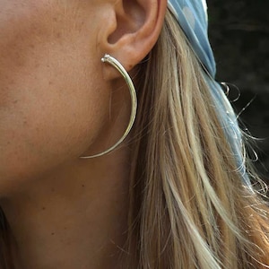 Op de afbeelding: Een zilveren oorring met een gebogen, puntig ontwerp. De oorring wordt gedragen in het linker oor van een persoon met lang blond haar.